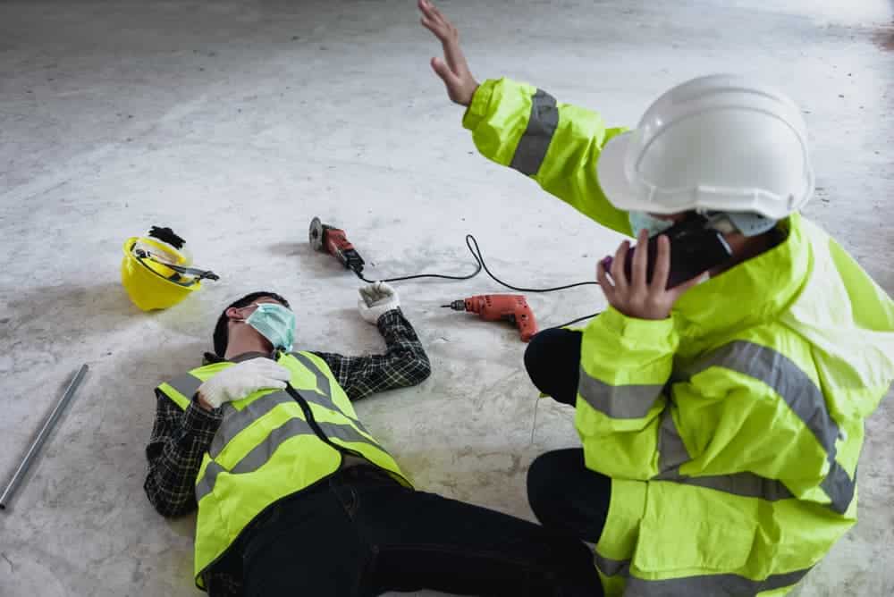 A worker experiencing a work accident at a construction site, shown unconscious while a colleague signals for help and calls the safety officer.