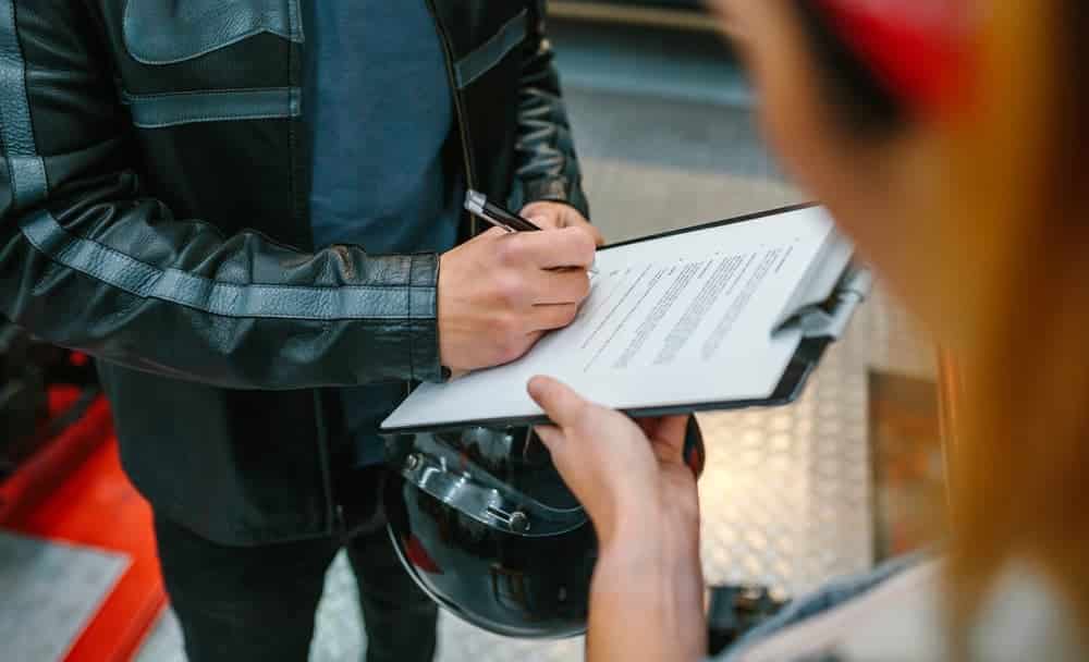 biker man wearing leather jacket and holding helmet signing insurance policy to receipt his repaired motorcycle on workshop