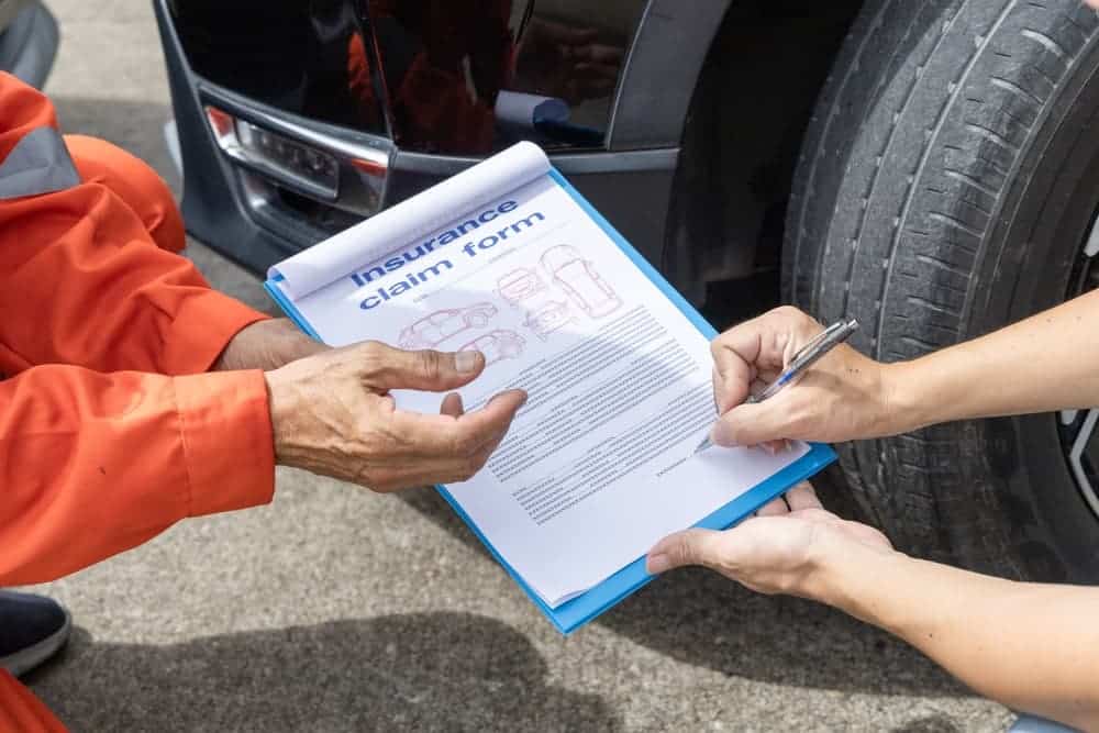 An insurance agent examines a damaged car after a road accident, while the car owner and insurance staff complete the necessary paperwork.
