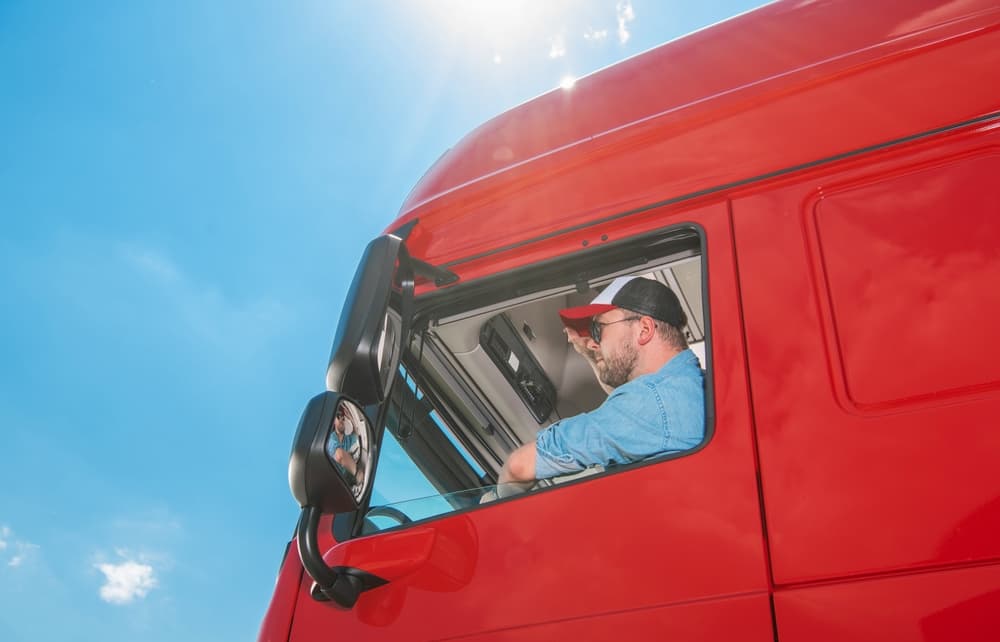 Truck driver in a blue shirt and cap sits in his large red lorry, looking ahead. A bright blue sky serves as the backdrop. Transportation concept.