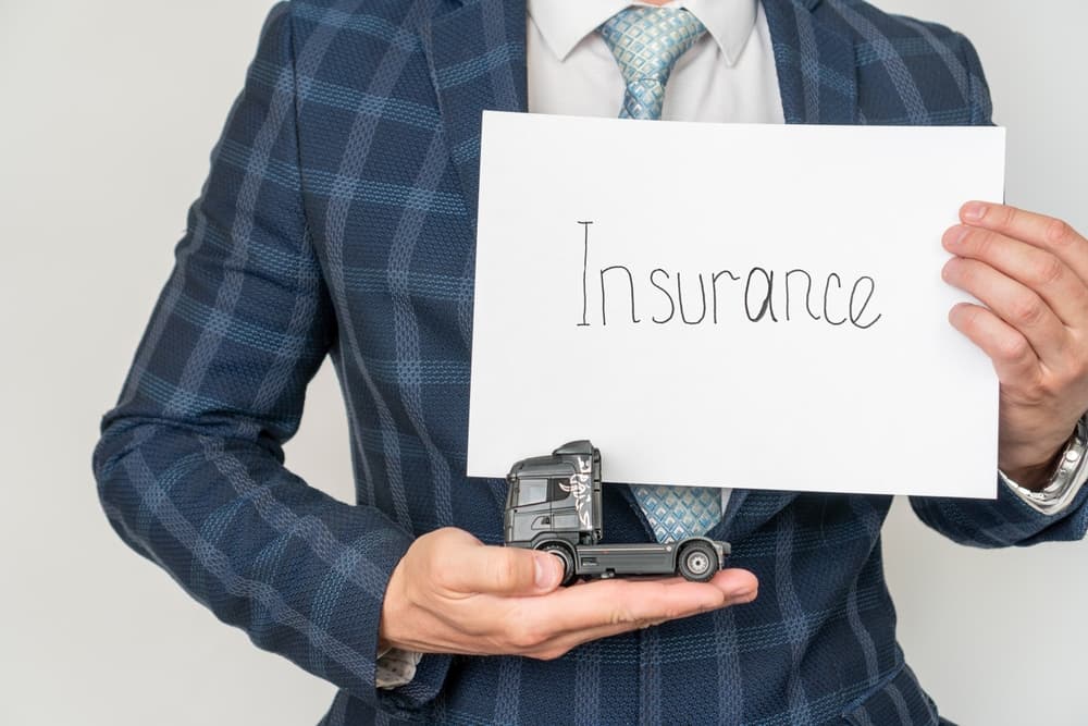 A businessman's hand shields a toy truck, symbolizing truck insurance. The poster features the word "Insurance," emphasizing protection and coverage.